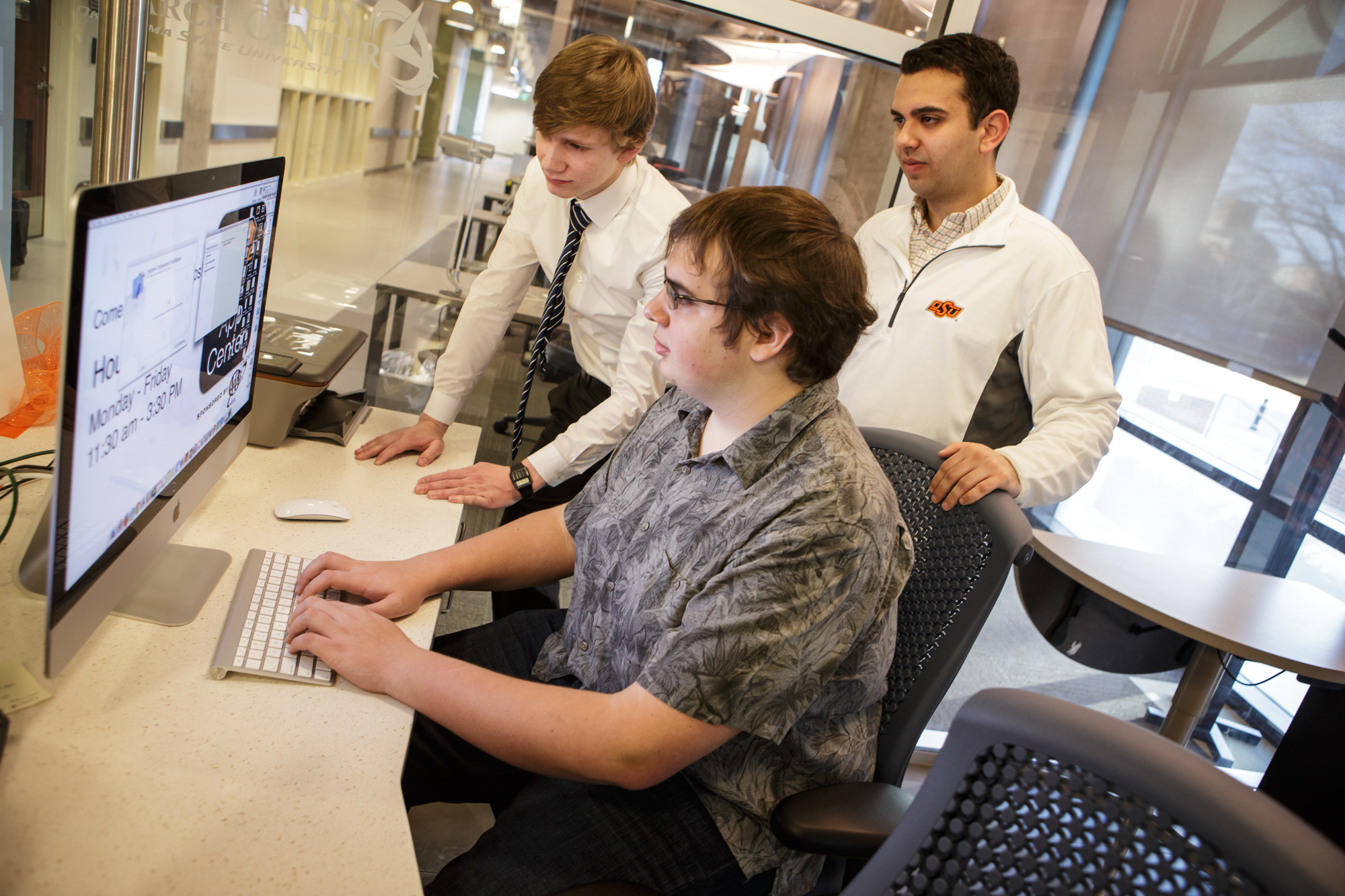 Three individuals in a modern office setting; one seated at a desk using a computer while two others stand nearby observing. 