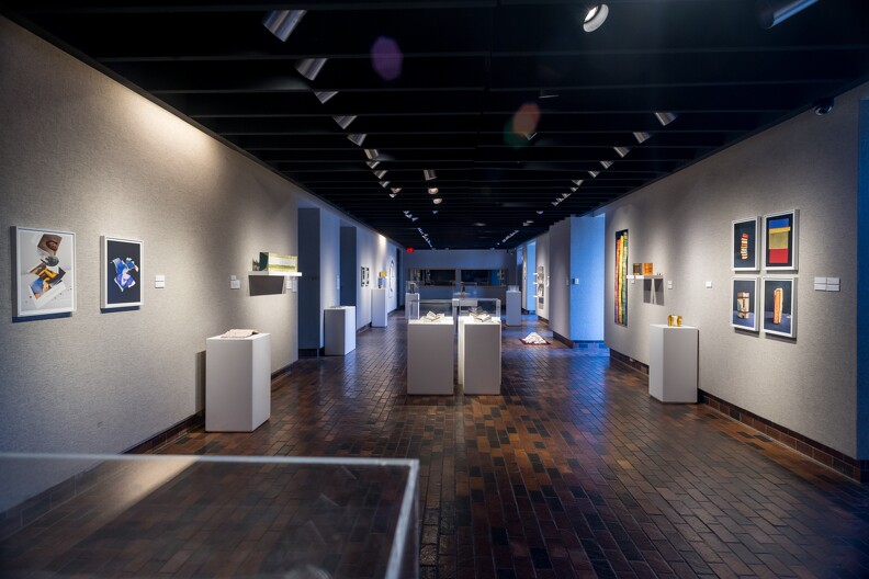 Interior of the Stillwater Museum of Art with dark brown wood floors, white walls. Colorful artwork hangs from the walls. Items are in white pedestals under display cases.