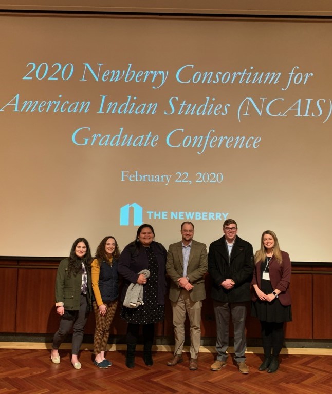 Four students and Dr. Boles stand in front of a protector screen with the Newberry logo behing them.