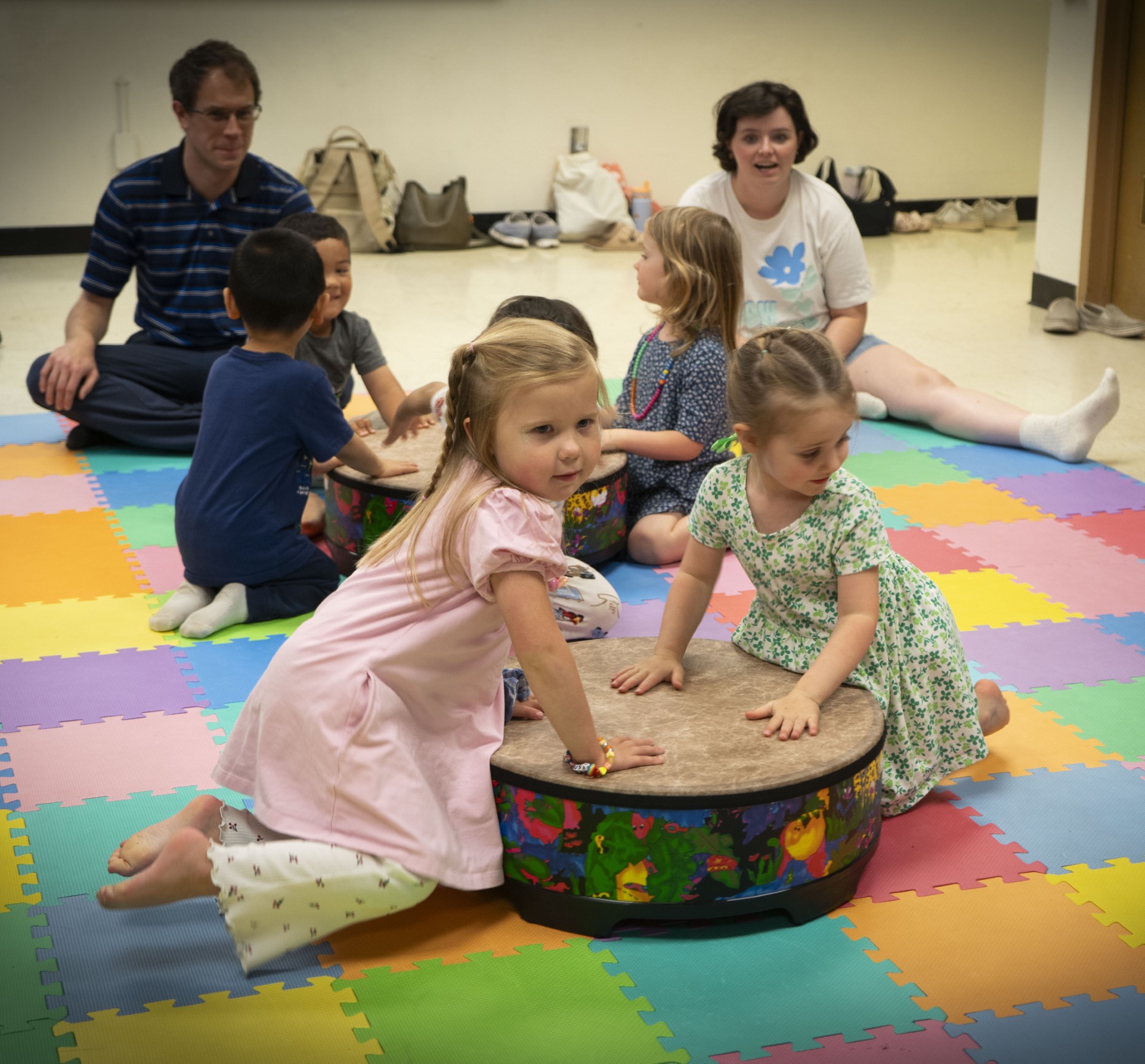 Two children playing drums with parents and other toddlers in the background.