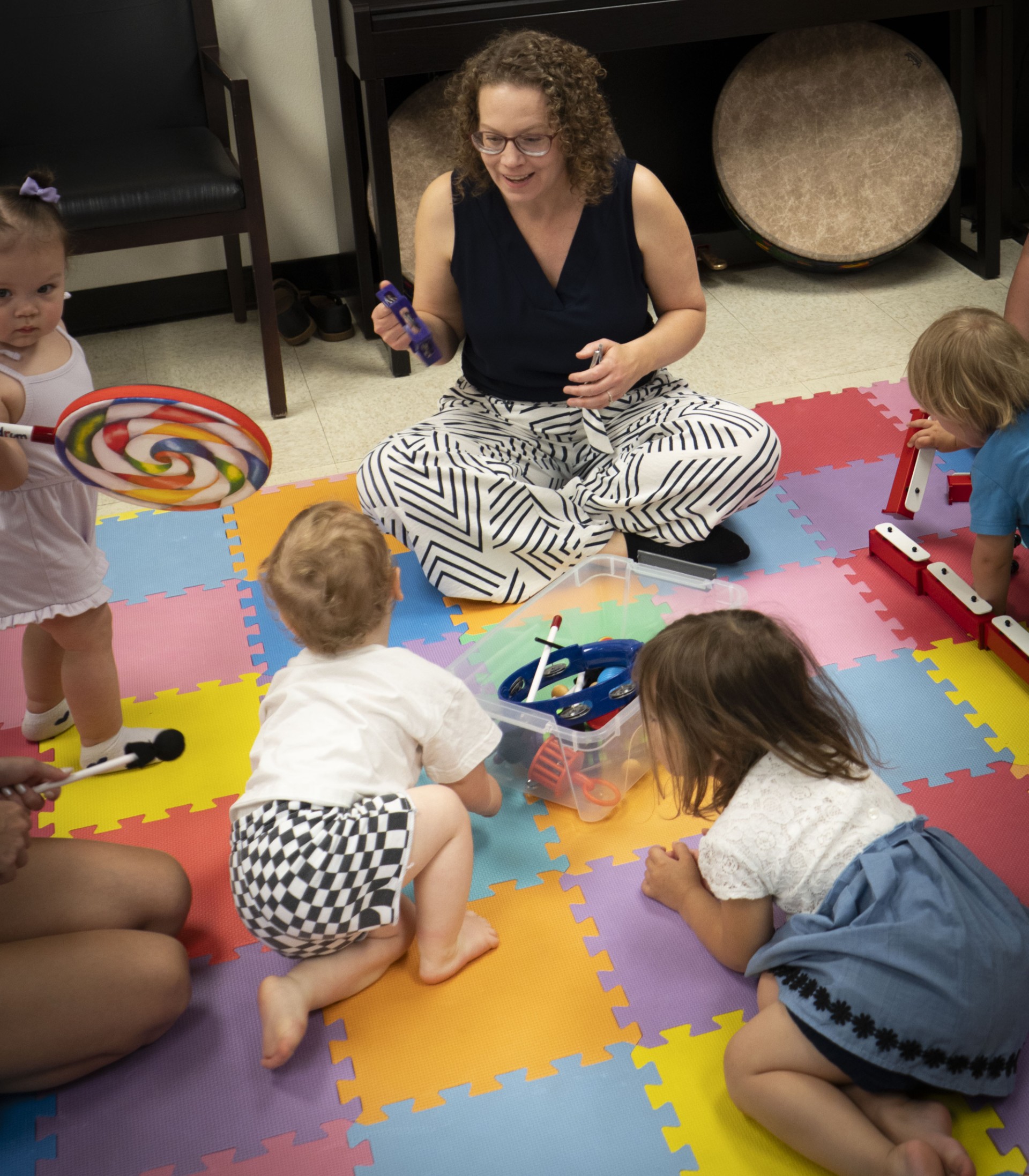 Music teachers sitting on a colorful floor with toddlers, each child holding a musical instrument during a group activity.