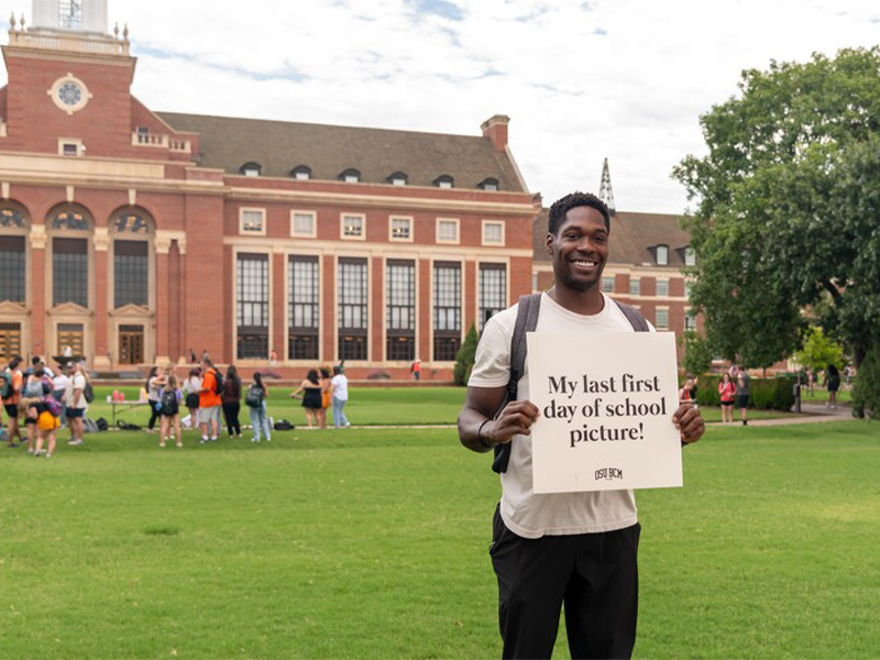A male student holding a sign that reads, 'This is my last first day of school' with a big smile, standing in front of a school building. A male student holding a sign that reads, 'This is my last first day of school' with a big smile, standing in front of a school building.